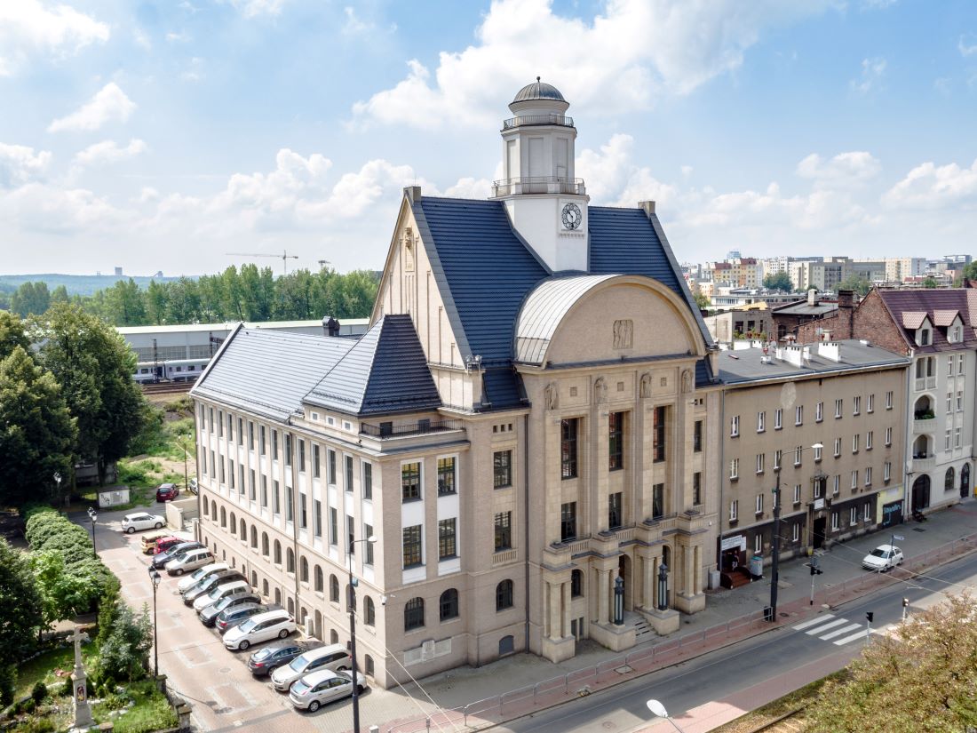 Daytime view of the historic main building of the University of Economics in Katowice (UEKat), a beige, neoclassical-style structure with tall arched windows, columns, and a distinctive central dome topped with a clock tower. The building has a dark slate roof with dormer windows and is situated along a city street with parked cars and surrounding residential buildings. In the background, green trees and the skyline of Katowice are visible under a partly cloudy blue sky.