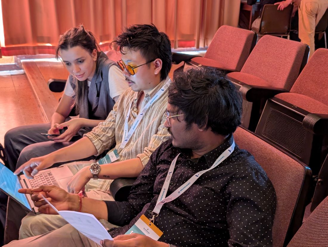 Three students are sitting in a confeence room next to each other. They are looking and pointing at a laptop screen that the student in the middle is holding.