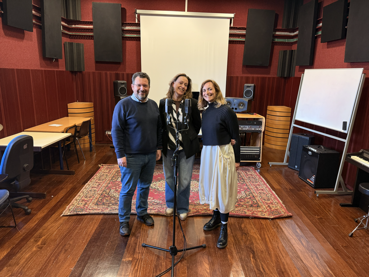 A man and two women are standing in front of a microphone in a room with sound proof panels and a big carpet.