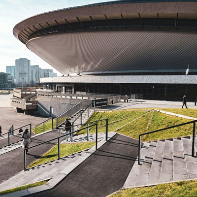 A panoramic view of the Spodek Arena in Katowice, Poland, at daytime. The iconic, UFO-shaped building has a circular structure. In the foreground, there is stairs leading up a green hill to the right. The sky above is in a bright blue colour and there are high-rise residential buildings in the background.