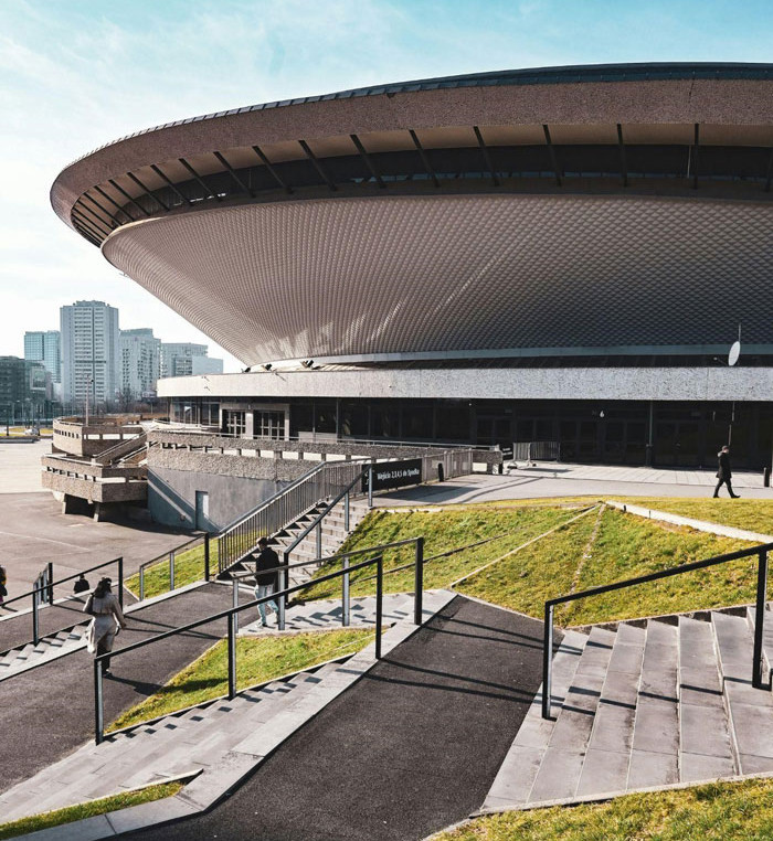 A panoramic view of the Spodek Arena in Katowice, Poland, at daytime. The iconic, UFO-shaped building has a circular structure. In the foreground, there is stairs leading up a green hill to the right. The sky above is in a bright blue colour and there are high-rise residential buildings in the background.