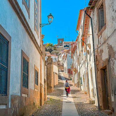 A person walks up a steep cobblestone street lined with historic buildings toward a castle tower under a bright blue sky.