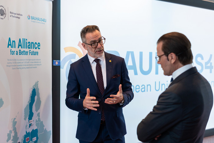 Two men in formal attire speak during a BAUHAUS4EU event, with the alliance logo and presentation screen visible in the background.