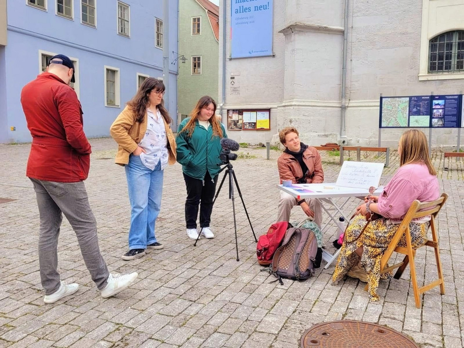 Group of five people outdoors with two seated at a table with papers and three standing nearby including one operating a camera on a tripod