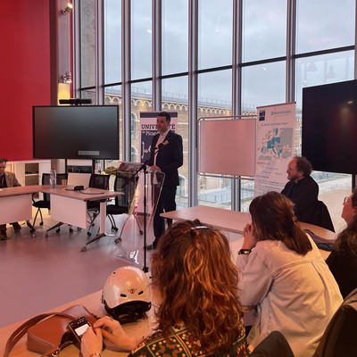 Prof. Peter Benz speaks at a transparent lectern during a meeting at Université de Picardie Jules Verne. Participants are seated around tables in a modern room with red walls, large floor-to-ceiling windows, and presentation screens. A BAUHAUS4EU banner and a whiteboard stand beside the speaker.