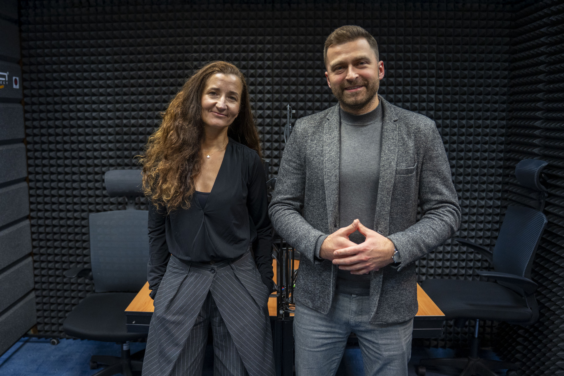 Two people standing in a room with soundproofing foam on the walls, one wearing a black top and striped pants, the other wearing a grey blazer and jeans