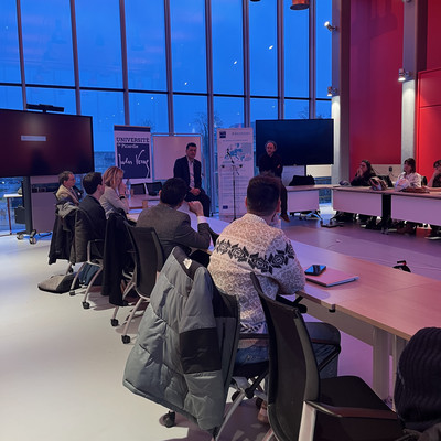 Prof. Peter Benz speaks at a transparent lectern during a meeting at Université de Picardie Jules Verne. Participants are seated around tables in a modern room with red walls, large floor-to-ceiling windows, and presentation screens. A BAUHAUS4EU banner and a whiteboard stand beside the speaker.