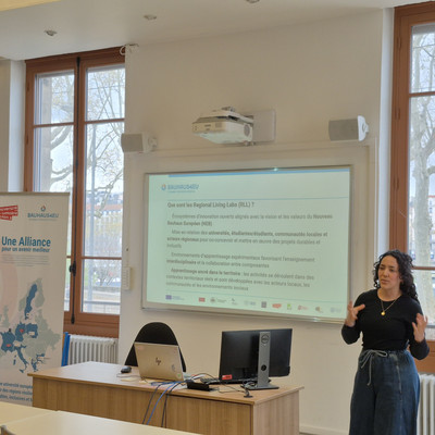 A presenter gives a BAUHAUS4EU presentation in a bright room with large windows.