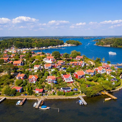 The red wooden houses of the Brändaholm allotments gardens on the peninsula Dragsö on a sunny day. There are many wooden jettys and a boat on the blue ocean.