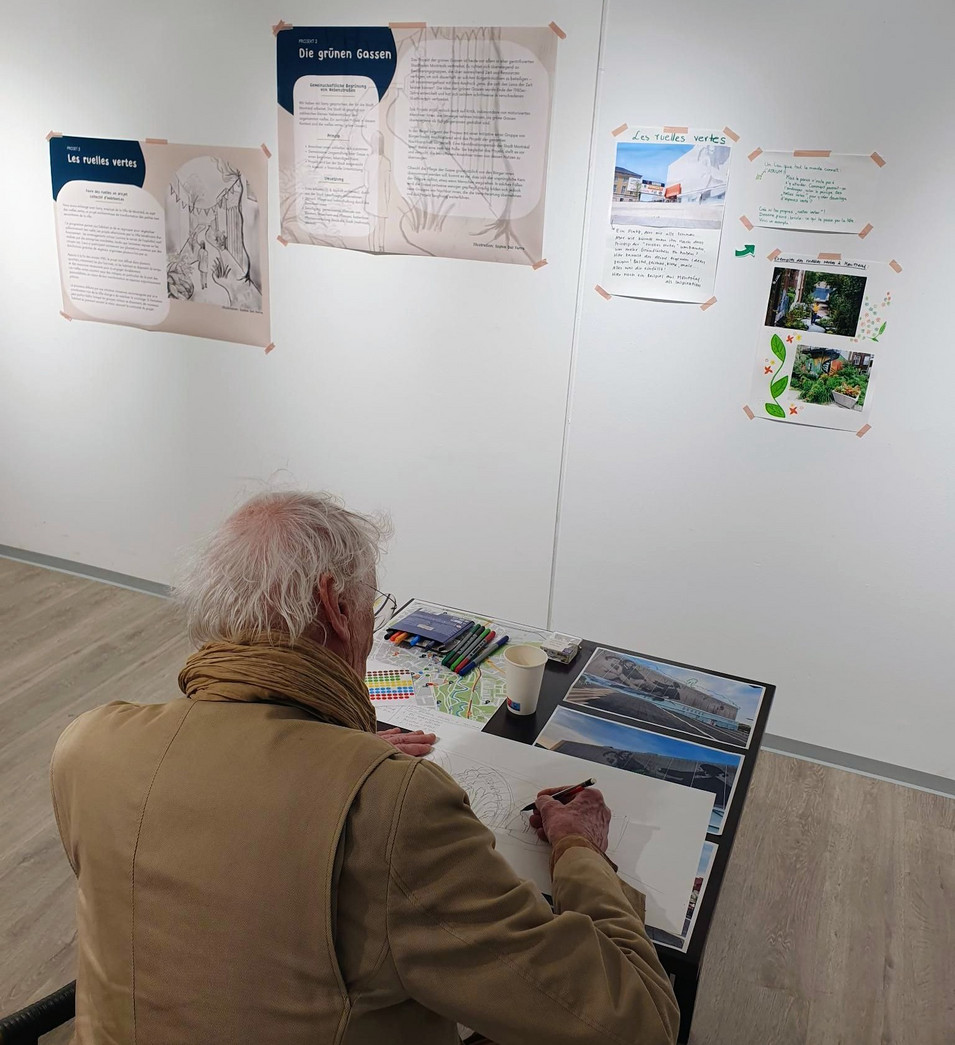 Older man with white hair and a brown scarf drawing at a table in a gallery with informational posters on the wall