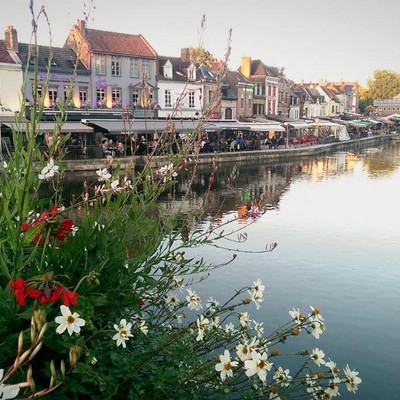 View of the Quai Saint-Leu in Amiens, with colorful flowers in the foreground, the Somme River in the middle, and the Quai Bélu with its buildings in the background