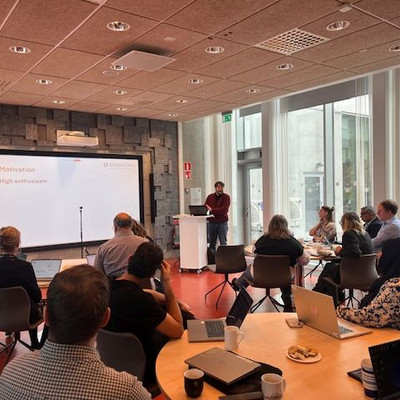 A speaker addresses participants seated in a conference room during a presentation.