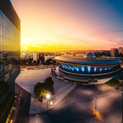 A panoramic view of the Spodek Arena in Katowice, Poland, captured at sunset. The iconic, UFO-shaped building is illuminated with blue lights around its circular structure. In the foreground, there is an open square with a few trees and streetlights, while reflections of the sunset and cityscape appear on the glass façade of a modern office building on the left. The horizon glows with warm orange and yellow tones, contrasting with the deep blue sky above and high-rise residential buildings in the background.