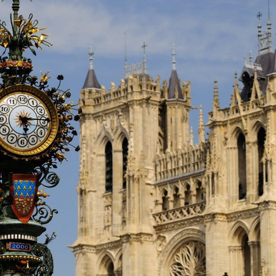 An ornate standing clock in front of the Cathédrale Notre-Dame d'Amiens, a cathedral in Gothic style.