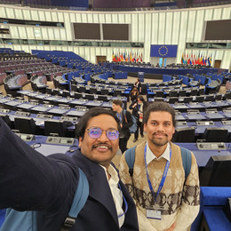 Two people taking a selfie inside the large circular parliamentary chamber of the European Parliament with rows of seats and flags behind a central podium