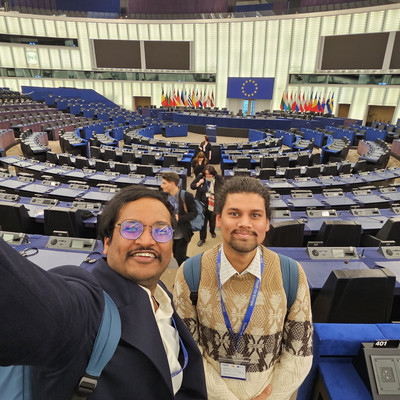 Two people taking a selfie inside the large circular parliamentary chamber of the European Parliament with rows of seats and flags behind a central podium